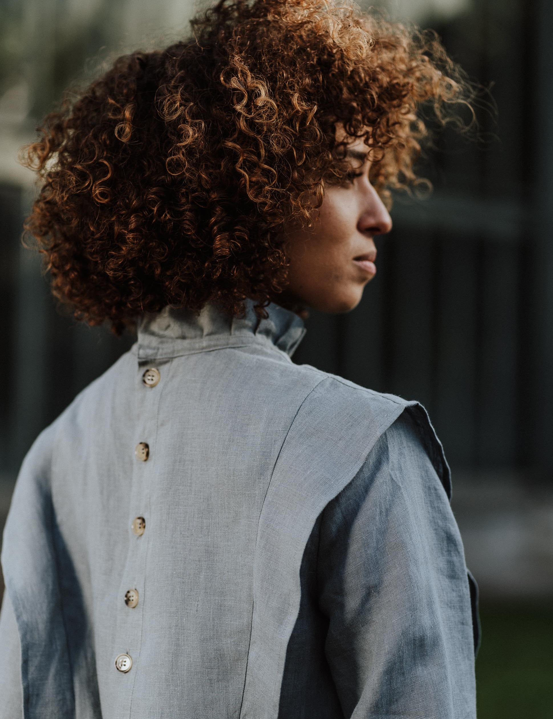 Rückansicht einer Frau, die die Margaret-Bluse in einer grünen Serre trägt. Die Bluse aus atmungsaktivem Leinen kombiniert Eleganz mit einem zeitlosen Design.