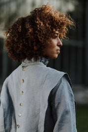 Rückansicht einer Frau, die die Margaret-Bluse in einer grünen Serre trägt. Die Bluse aus atmungsaktivem Leinen kombiniert Eleganz mit einem zeitlosen Design.