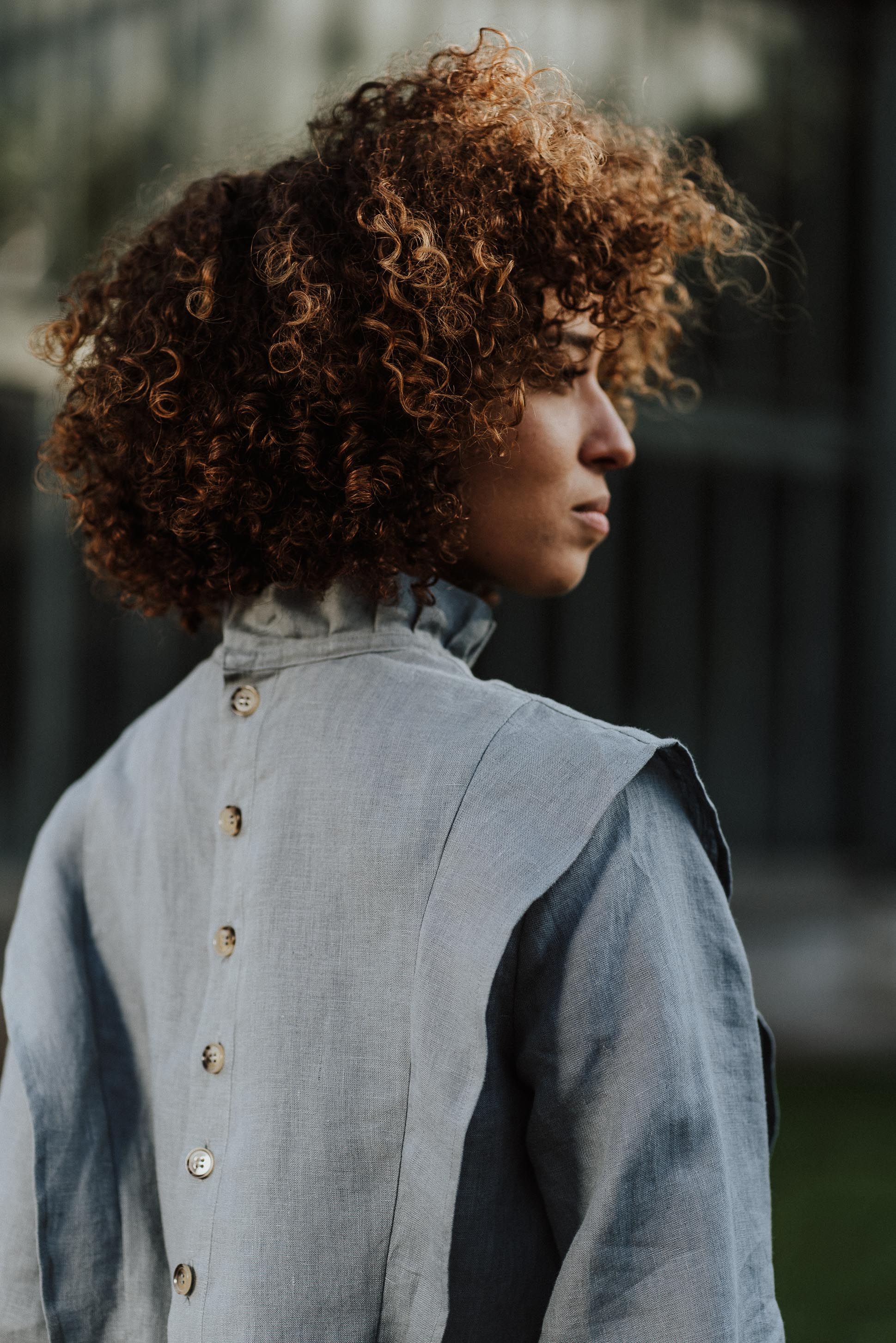 Rückansicht einer Frau, die die Margaret-Bluse in einer grünen Serre trägt. Die Bluse aus atmungsaktivem Leinen kombiniert Eleganz mit einem zeitlosen Design.