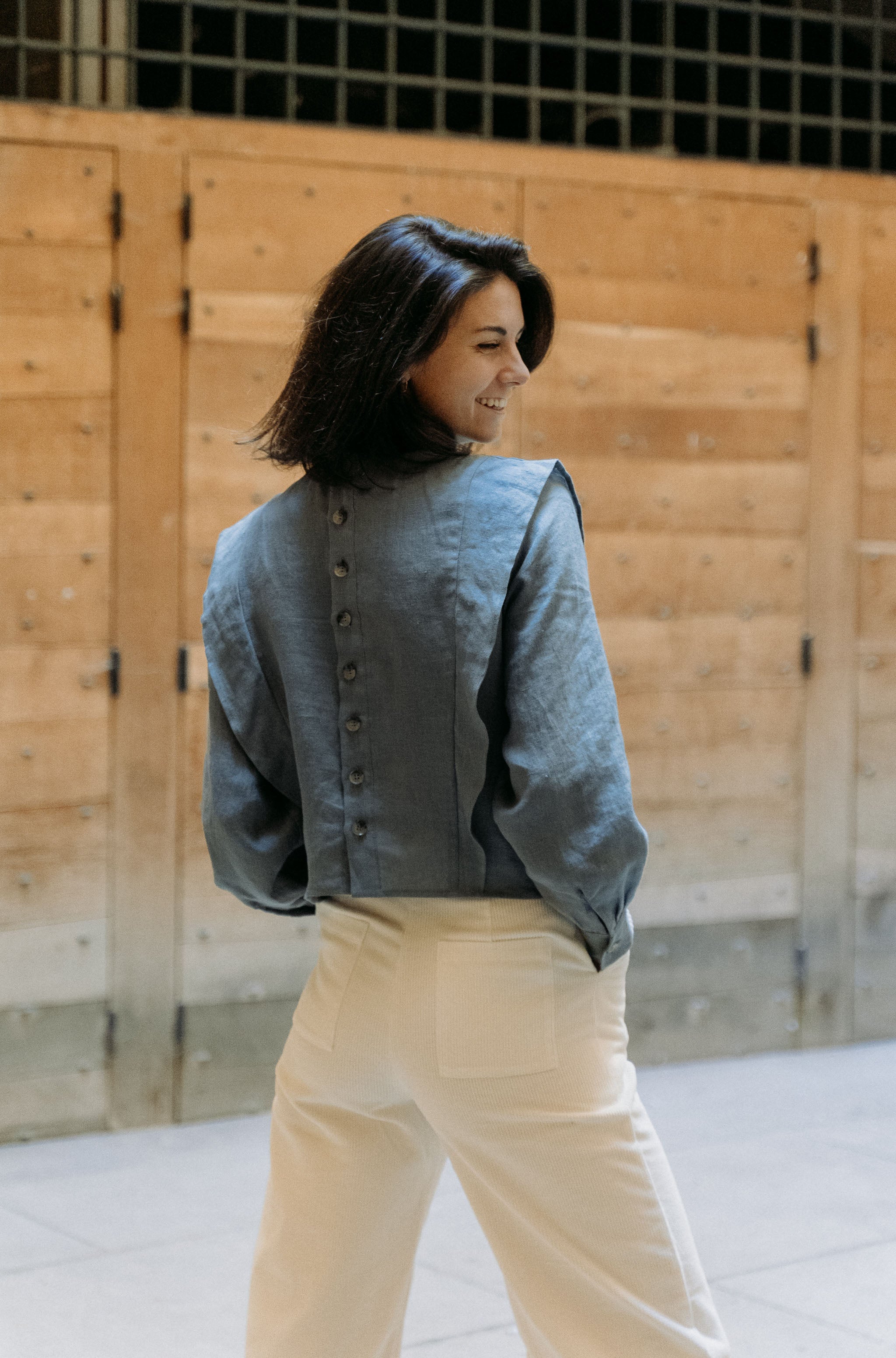 Eine Frau trägt die Margaret-Bluse aus schieferblauem Leinen und zeigt die Knopfleiste auf der Rückseite. Ein elegantes und vielseitiges Design für stilvolle Looks.