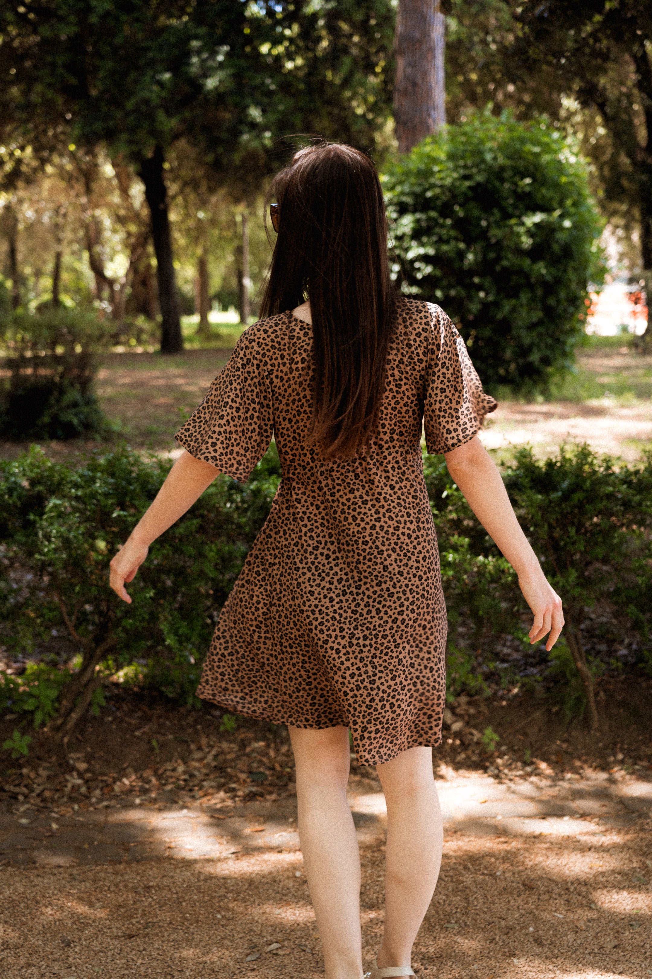 Frau im Sommerkleid mit Leo-Print von hinten, elegant im Park – nachhaltige EcoVero™-Viskose, eleganter Schnitt und femininer V-Ausschnitt für zeitlose Mode.
