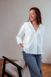 Frau trägt die weiße Stillbluse Camille im Innenraum, steht aufrecht und zeigt die elegante, gerade Passform und die 3/4-Ärmel des Modells.