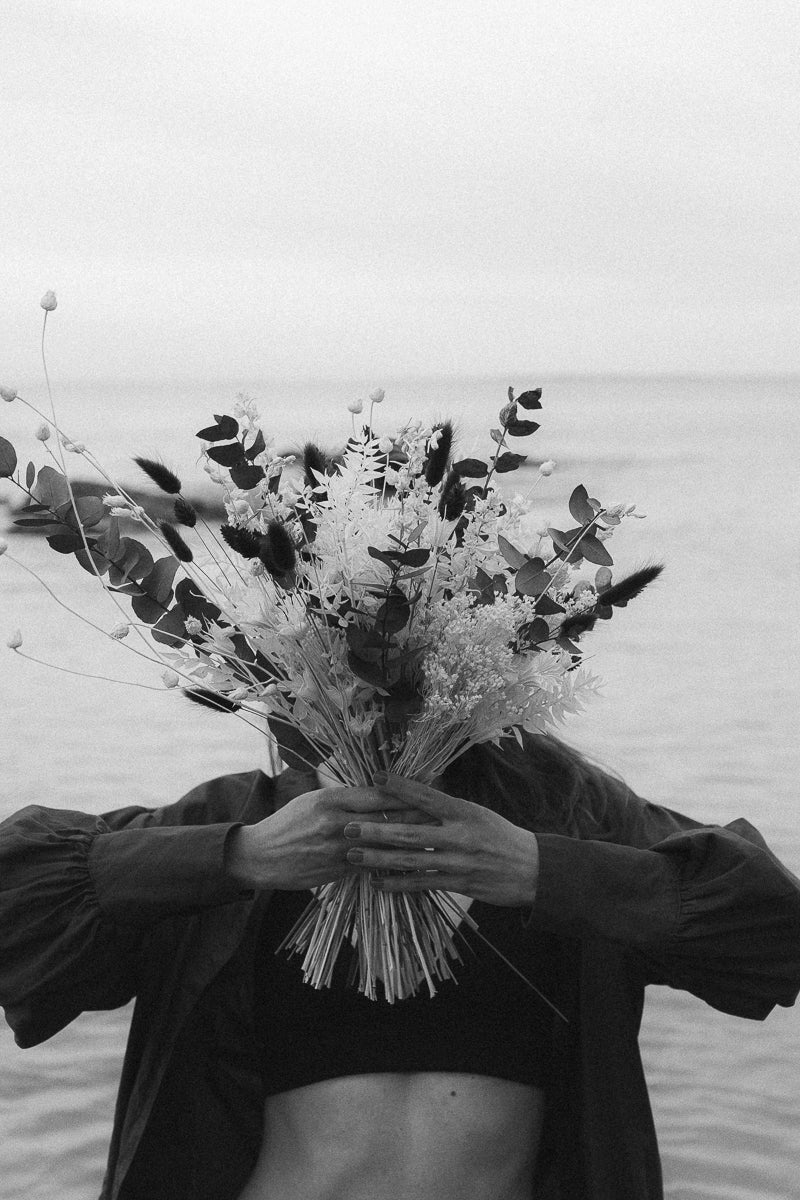Schwarz-Weiß-Foto einer Frau am Strand, die einen Blumenstrauß vor ihrem Gesicht hält, was eine Atmosphäre von Ruhe und Natur vermittelt.