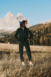 Frau trägt den grünen Baumwollcord-Einteiler Dolomites mit minimalistischer Schnittführung, umgeben von einer beeindruckenden Berglandschaft.