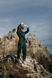 Frau lacht und trägt den Einteiler Dolomites aus grünem Cordstoff, während sie auf einem Felsvorsprung steht, mit weitem Blick auf die Berge.