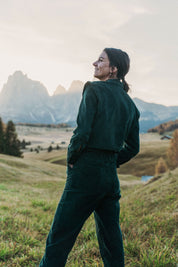 Frau steht mit dem Rücken zur Kamera, trägt den Einteiler Dolomites aus grünem Baumwollcord in einer Berglandschaft.