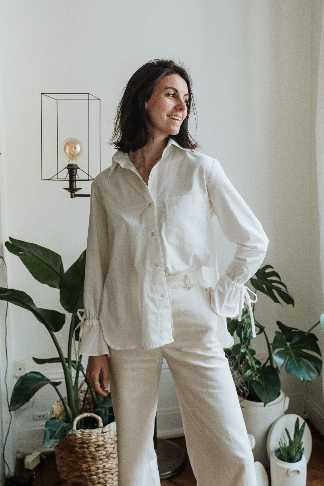 Eine Frau trägt die weiße Noosa-Bluse in einem minimalistischen Innenraum. Die Bluse besticht durch ihren zeitlosen Stil und die edlen Details.
