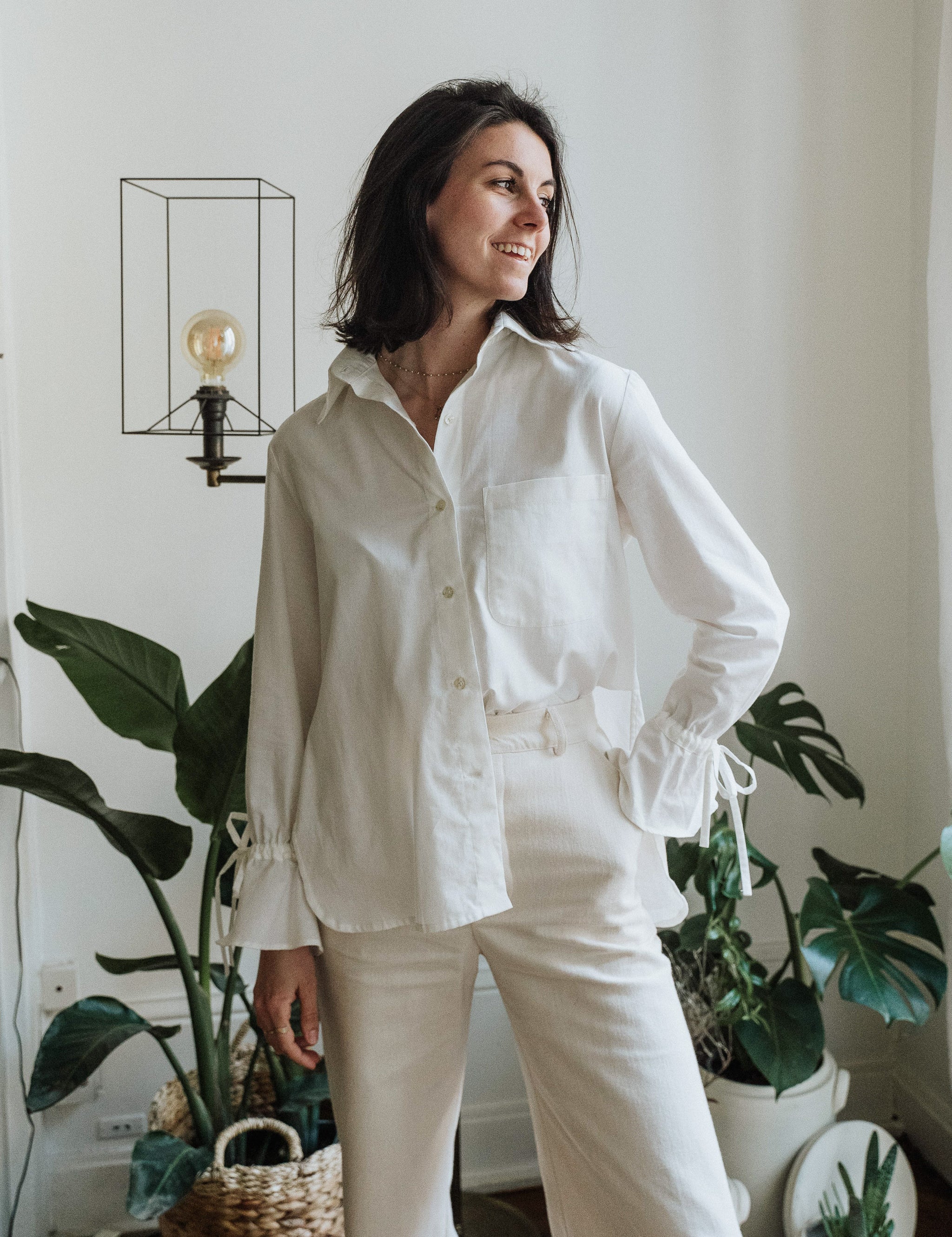 Eine Frau trägt die weiße Noosa-Bluse in einem minimalistischen Innenraum. Die Bluse besticht durch ihren zeitlosen Stil und die edlen Details.