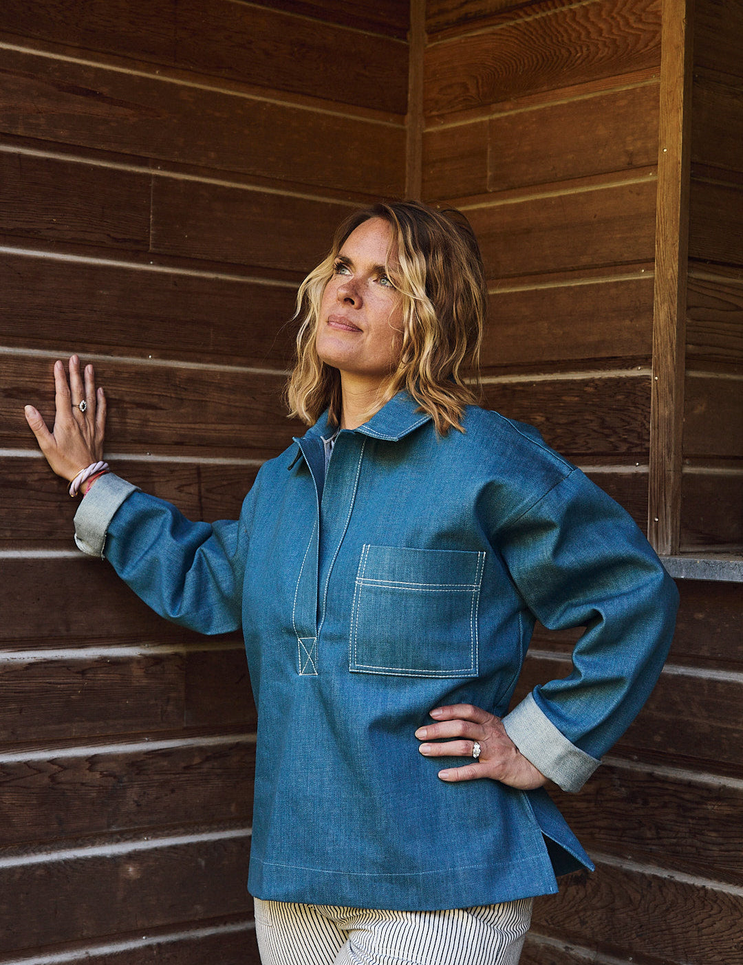Frau steht draußen und trägt eine lockere Bluse im Seglerstil aus blauem Baumwoll-Denim, hüftlange Passform, klassische Streifenoptik