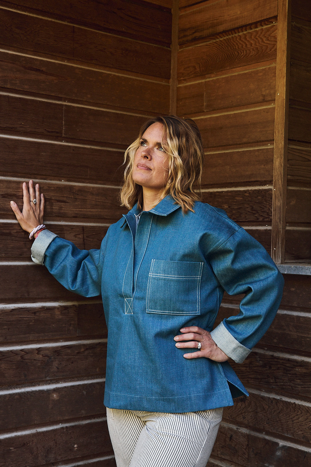Frau steht draußen und trägt eine lockere Bluse im Seglerstil aus blauem Baumwoll-Denim, hüftlange Passform, klassische Streifenoptik