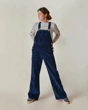 Eine Frau trägt eine blaue Cord-Latzhose, kombiniert mit einem gestreiften Langarmshirt aus 100 % Bio-Baumwolle. Hergestellt in Frankreich, fair produziert und nachhaltig – klassische französische Damenmode für Alltag und Büro.